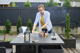 A man is arranging a fresh salad on a table in an inviting garden area. The sun is shining