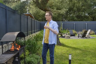 A man stands next to a barbecue grill, casually holding a drink while readying food. The scene is