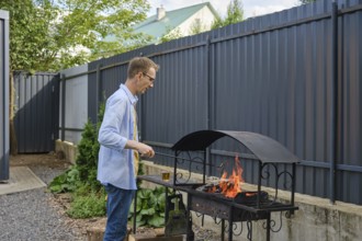 A man stands by a grill in a backyard, tending to the flames. The surrounding area features