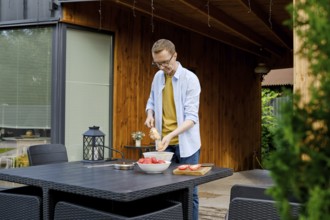 A man is joyfully preparing a meal outdoors at a wooden kitchen area. He strands meat on a skewer