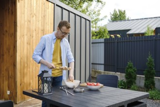 A man wearing a yellow shirt and denim jacket is preparing meat skewers at a wooden table in a