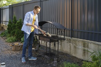 A middle-aged a man puts firewood in the grill in a backyard. He is going to cook meat outdoors on