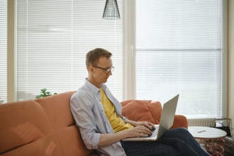 A retired man sits comfortably on a couch and writes his memoirs on laptop in a well-lit living