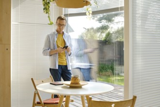 A middle-aged man stands in a bright, modern kitchen checking weather forecast at his smartphone.