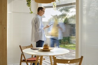 A man stands inside a stylish room, using his smartphone while looking out through a large glass