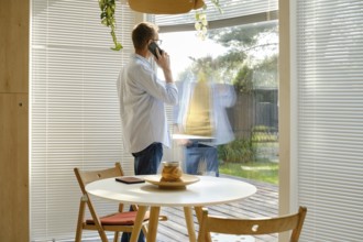 A middle-aged man stands near a large window, engaged in a phone conversation. Sunlight filters