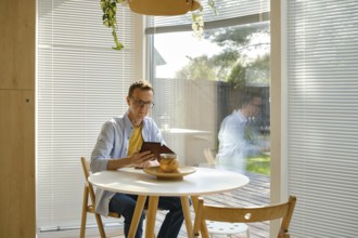 A man sits at a round wooden table in a modern kitchen, focused on his e-book. Sunlight filters