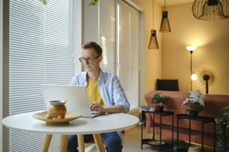 A freelancer is typing on a laptop at a round table in a modern home workspace. Natural light pours