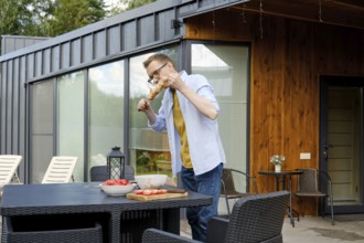 A man is enjoying a summer barbecue at a stylish wooden house. He is focused on grilling meat,
