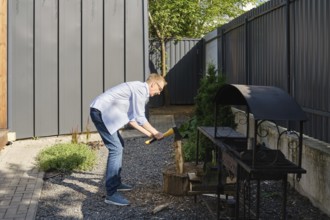 A middle-aged man is chopping a piece of wood in a well-maintained backyard garden. He is dressed