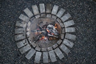 Bright flames dance among burning logs in a circular stone fire pit during early evening. The warm