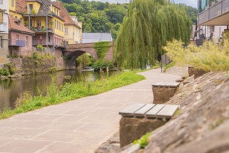Paved path along a river with stone wall and benches, a bridge in the background, small town of