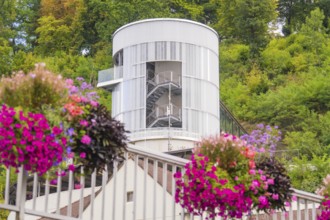 White tower with stairs surrounded by green trees, colourful flowers in the foreground, small town
