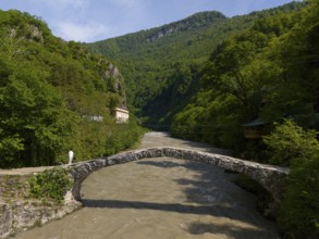 Person crossing a stone bridge over a river, surrounded by green hills and lush forest, aerial