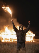 Maskierte Person hebt Fackel vor brennendem Strohlager in der Nacht, Brandstiftung, Brandleger,