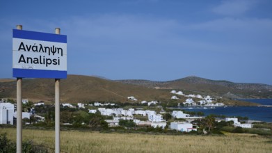 A town sign in front of a coastal settlement with whitewashed houses and blue sky, Analipsi,