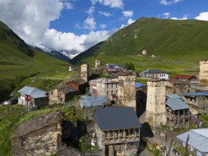 Medieval village with stone towers in a green valley surrounded by mountains and cloudy sky, aerial