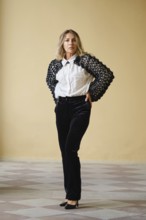 A middle-aged woman stands confidently with her hands on her hips, wearing a fashionable black and