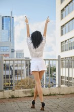A woman stands outdoors in a vibrant urban setting with arms lifted to the sky. Dressed in a