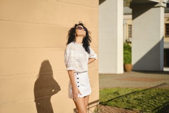 A woman stands playfully against a tan wall in a bright outdoor environment, enjoying the warm sun.