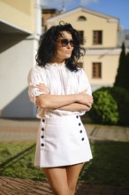 A woman with dark curly hair poses with crossed arms in a fashionable white outfit featuring a