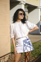 A woman with curly hair stands confidently in a fashionable outfit featuring a white blouse and a