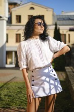 Bright sunlight highlights a woman posing confidently in a chic white blouse and skirt. She stands