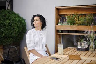 A woman with curly black hair sits at a wooden table in an outdoor cafe, gazing thoughtfully in a