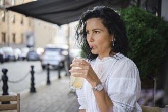 A woman sits at a sidewalk cafe, sipping a cold lemonade through a straw. She wears a white blouse