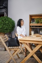 A woman with curly hair relaxes at a wooden table in an outdoor cafe, dressed in a fashionable