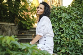A woman with curly hair is seated gracefully on stone steps surrounded by vibrant green plants. She