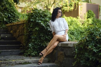 A woman with curly dark hair and glasses sits on a stone wall in a garden, surrounded by greenery.