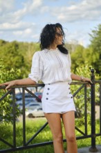 In a vibrant park setting, a woman poses confidently in a white blouse and mini skirt. Sunlight