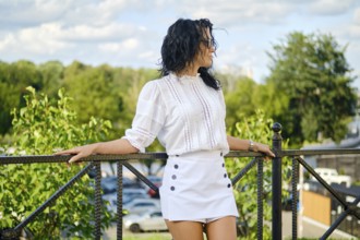 A woman with curly hair stands on a balcony, leaning against a railing. She wears a white blouse
