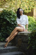 A woman sits elegantly on a stone wall in a vibrant garden, wearing a chic outfit and sunglasses.