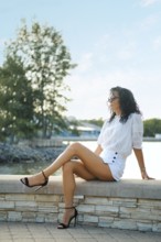 A woman with curly hair relaxes on a stone bench by the water, dressed in a chic white outfit and