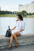 A fashionable woman sits by the water, dressed in a white top and shorts while wearing stylish