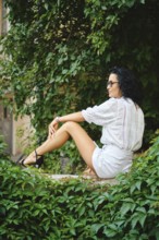 A woman with dark hair sits peacefully in an urban garden surrounded by vibrant green leaves. She