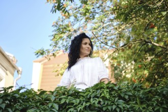 A woman with dark, curly hair stands amidst lush green leaves, looking thoughtfully into the