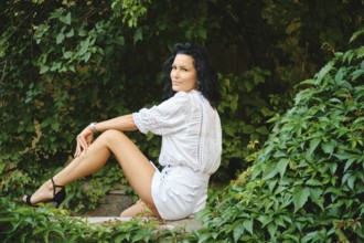 A woman in white blouse and shorts sits relaxed in a vibrant garden, surrounded by greenery