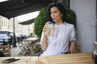 A woman with curly black hair sits at a cafe terrace enjoying a drink. She is wearing a stylish