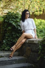 A woman sits elegantly on a stone ledge in a garden, dressed in a stylish white top and mini skirt.