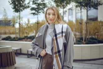 A young woman stands outdoors, showing off her fashionable gray coat featuring a plaid pattern. She