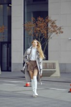 A confident woman strolls along a city street, wearing a trendy autumn outfit that includes boots