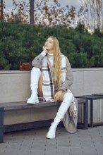 A dreamy woman sits thoughtfully on a bench in a park, dressed in a fashionable outfit with a long