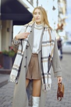 A fashionable young woman strolls down a cobblestone street, wearing a long coat and a plaid scarf,