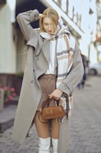 Stylish woman in beige coat and white boots walking down the street stopped to adjust her scarf