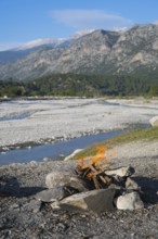 A campfire in a rocky landscape with mountains and a riverbed in the background, Dargaz River near