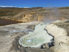 A geothermal area with a steaming geyser and rocks in a wild landscape under a blue sky, aerial