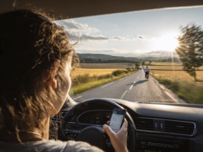 A car driver is dazzled by the sun and looks at her smartphone while driving, putting herself, the